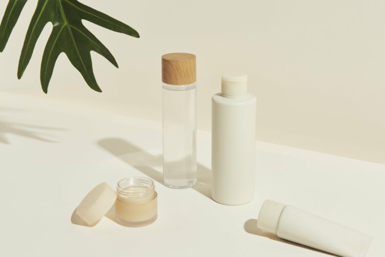 Verschiedene kosmetische Flaschen und Tiegel, darunter ein Glas mit Holzstoß, auf einem hellen Untergrund.