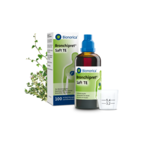 Bronchipret Saft TE in einer braunen Flasche, mit grüner Verpackung und einem Messbecher daneben, umgeben von Kräutern.